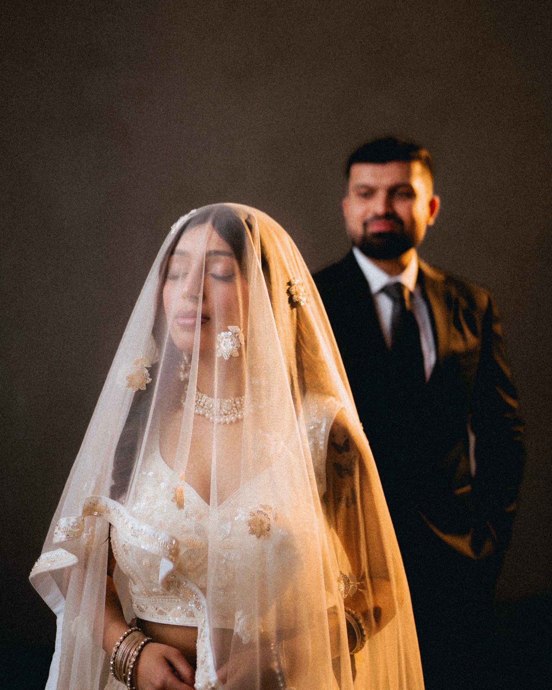 A bride under a long sheer veil beside her groom against a dark backdrop