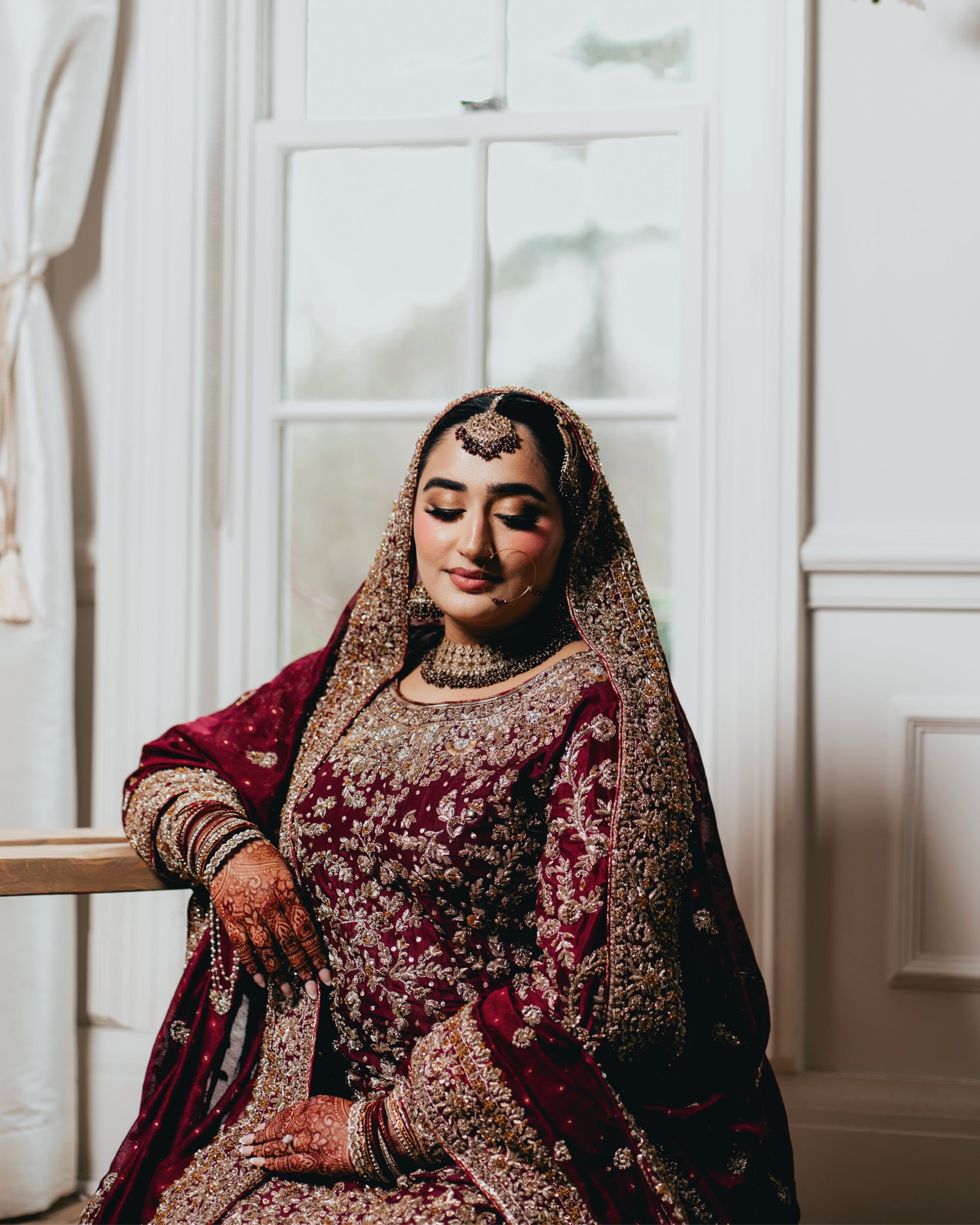 A bride in a maroon lehenga sits in soft window light in a white panelled room